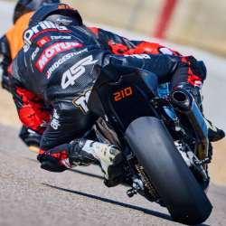 Rider in black and red 4SR leathers on a black Aprilia motorcycle numbered 210; seen from behind with more of the red and black leathers exposed and orange material visible at the shoulder; number 210 is visible on the tail