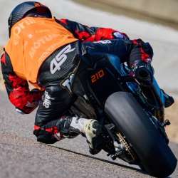 Rider in black and red 4SR leathers on a black Aprilia motorcycle numbered 210; seen from behind wearing an orange vest over the leathers; number 210 remains visible on the tail
