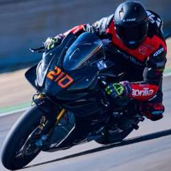 Rider in black and red 4SR leathers on a black Aprilia motorcycle numbered 210; seen from a front three-quarter angle on the black motorcycle numbered 210