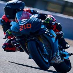 Rider in black and red 4SR leathers on a black Aprilia motorcycle numbered 210; seen from the front on the black motorcycle with number 210 visible