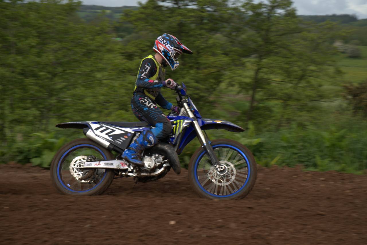 A motocross rider on a blue motorcycle passes trees on a dirt course.